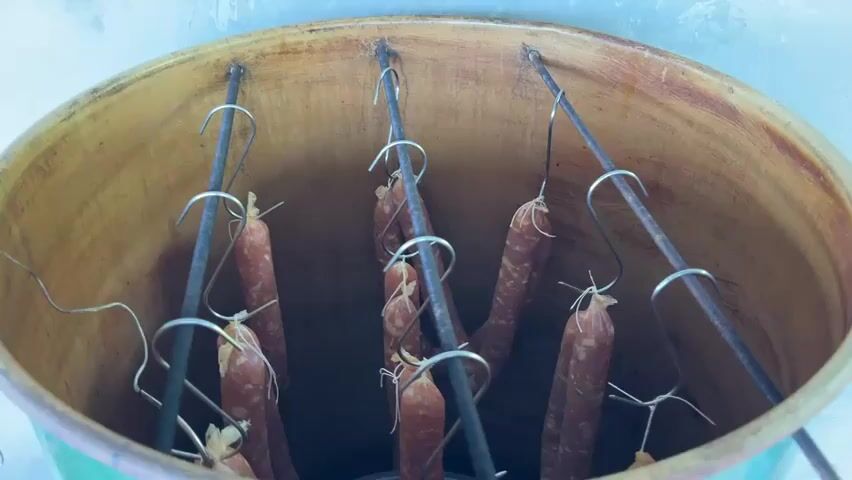 The video shows a close-up, top-down view inside a smoking chamber with several strings of uncooked sausages hanging from metal rods by hooks. The sausages are reddish-brown and appear to be dry-cured or cured before smoking. The interior of the chamber is made of wood or a similar material, and the sausages are suspended in the center.