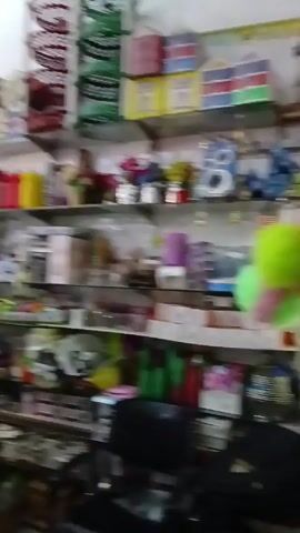 The video shows the inside of a crowded shop, filled with an assortment of household items and decorations. Several people, including women wearing hijabs and children, are browsing and interacting near the shop's checkout area.  Shelves lining the walls are stacked with various products, including plastic containers, decorative items, and packaged goods. Signs with the price "$1$" are visible above some of the merchandise. The lighting is somewhat dim, creating a sense of a bustling, cramped retail environment.