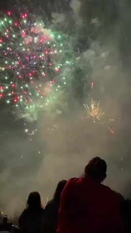 A short video clip shows a fireworks display at night. The sky is filled with bursts of light in various colors, including white, gold, green, red, and purple. Smoke from the fireworks drifts through the air. In the foreground, silhouetted figures of people are visible watching the display. Some of the fireworks are in focus, while others are blurred. The overall scene is dark, with the bright lights of the fireworks being the primary source of illumination.
