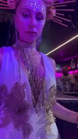A woman with glitter on her face and chest, wearing a white dress with gold embellishments and a floral headpiece, dances under purple lights. She has a long, beaded necklace. The background appears to be a nightclub or bar with other people visible.