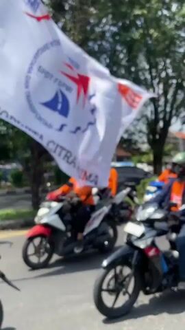The video shows a daytime procession of people riding motorcycles on a sunny street, likely in Tebel, Indonesia. Most participants are wearing orange or blue shirts and helmets. A large white flag is prominently displayed, featuring the text "FSPMI" and "PUK. BUKAT," along with the logo of the Federasi Serikat Pekerja Metal Indonesia (FSPMI). A red and white Indonesian flag is also visible. The motorcyclists are driving past trees and buildings, suggesting an urban or semi-urban environment. One visible license plate begins with "H," consistent with Central Java, Indonesia.