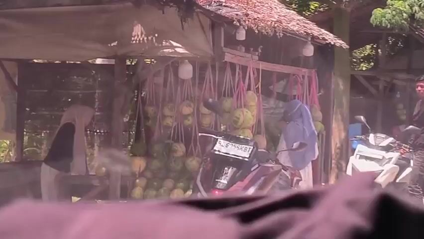 A man in a black jacket and a woman in a white hijab are near a roadside stand selling coconuts. The coconuts are hanging in bunches from the roof of the stand, which is made of wood and has a thatched roof. The man is standing next to a white scooter and looking at the coconuts. A poster is visible with a picture of a man on it. There are also blue bins next to the stand, and some fruits piled on top of them. The scene appears to be outside in a sunny, tropical environment. The camera seems to be inside a vehicle looking out.