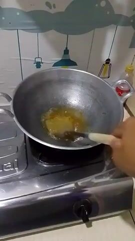 A person's hand is visible on the right side of the frame, holding a wooden utensil and stirring a light brown liquid in a wok. The wok is on a gas stove, with flames visible underneath. The background shows a kitchen wall with a white tile backsplash and a patterned border. A bottle of liquid and a can are on a shelf in the upper right corner.