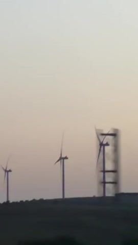 The video shows a blurry, low-angle shot of wind turbines against a pale sky. The turbines are silhouetted against the sky and appear to be in motion. The foreground is dark and indistinct, suggesting it might be a field or a road. The overall impression is of a vast, open landscape with renewable energy infrastructure.