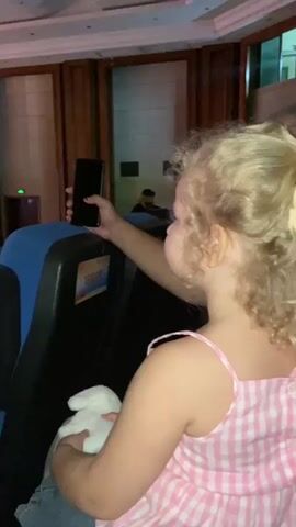 A young blonde girl with curly hair and a pink and white checkered dress is sitting in a dark room, possibly a theater or auditorium, holding a white stuffed animal and a smartphone. She is looking to her right and holding the phone up, as if taking a photo or video. The phone is dark, indicating the screen is off or displaying a black image. There are rows of empty blue and black seats in front of her. The room has wood-paneled walls and a high ceiling with lights. Other people are visible in the background, some seated and some standing. The overall lighting is dim and warm.
