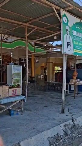 The video begins with a wide shot of a sign welcoming "SAMITO H. MUHAMMAD UMUR" from "MAKKAH AL-MUKARRAMAH & MADINAH AL-MUNAWWARAH" and wishing them a blessed Hajj. The sign features an image of the Kaaba. Beneath the sign, a covered structure appears to be a shop or stall. A table with various items, including what looks like religious texts and a statue, is visible. The ground is dirt and cracked concrete. The camera then pans to the right, revealing more stalls with clothing, including t-shirts and dresses, hanging for sale. A parrot decoration is also visible. The camera moves further to show a corrugated metal structure, possibly a wall or building. Finally, the video pans to an open area with trees and a clear blue sky. The overall impression is of a market or roadside stall area in a warm climate, possibly near a place of religious significance.