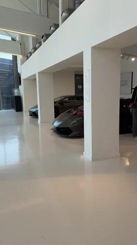 The video begins with a wide shot of a modern, minimalist showroom with a white floor and white columns. Several luxury sports cars are displayed, including a silver Lamborghini with red accents in the foreground. In the background, a black Lamborghini with yellow accents is visible, as well as another dark-colored sports car. Two people, a woman in dark pants and a man in a patterned shirt and shorts, are standing near the black and yellow Lamborghini, seemingly examining it. Another person, a child in a red shirt and shorts, is crouching down near the silver Lamborghini, looking at it. A second child, a girl in a white outfit, is also near the silver Lamborghini, holding up a phone or camera. The video pans slightly to the right, showing another section of the showroom with more cars and people. A man in a white t-shirt and blue shorts with tattoos on his arm is looking towards the cars. The overall impression is a well-lit and spacious showroom exhibiting high-performance vehicles.