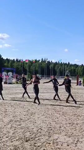 A group of five individuals, appearing to be women dressed in black outfits, perform a coordinated dance routine on a sandy equestrian arena. The individuals are spaced apart and moving in unison, executing various arm and leg movements. The background features a clear blue sky with scattered clouds, a line of trees, and flags mounted on poles. There are also structures visible in the background, possibly part of an event venue. The lighting suggests it is daytime, likely a sunny afternoon.