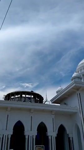The camera slowly pans upward, revealing the white walls and arches of a mosque building. A partial, dark wooden framework is visible to the left. The focus shifts to a large, white dome with supporting columns, and a sign reading "MASJID BESAR AL-FURQAN". The sky above is blue with scattered clouds.