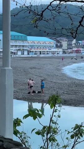 The video shows a beach with dark sand or pebbles.  In the foreground is a small pool of water reflecting the sky.  A tree with bare branches frames the top of the scene.  Behind the beach, there is a white building with a blue roof.  People are walking on the beach.  Three men are standing near the edge of the water, one wearing a blue shirt and two shirtless.  In the distance, there is a hillside.  The sky is overcast.  Waves are gently breaking on the shore.
