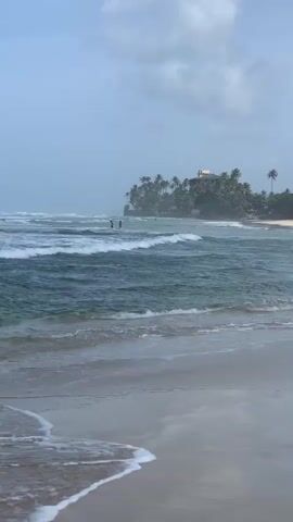 A wide shot of a beach on a cloudy day. Waves crash onto the shore in the foreground. In the mid-ground, a body of water is visible, with two people standing in the shallow water. In the background, a small island covered in palm trees is visible. A building sits atop the island. The shoreline to the right of the island is lined with palm trees and a few buildings. The sky is overcast.