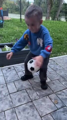 A young boy is playing with a soccer ball on a paved patio. He is wearing a blue sweatshirt, grey pants, and black shoes. The boy is in motion, appearing to be kicking or dribbling the ball. In the background, there is green grass, some plants, and a body of water. A trampoline and part of a fence are also visible.
