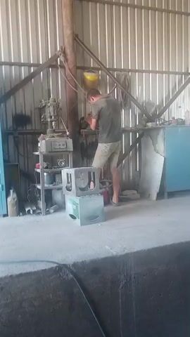 The video shows a man working in what appears to be a workshop or garage. He is wearing a dark t-shirt and light-colored shorts. He is standing and focused on a piece of machinery, which he is actively manipulating with his hands.

Several objects are visible in the scene. To the left of the man, there's a multi-tiered metal shelf unit with various items on it, including what looks like a drill press, some containers, and electrical components. In front of the shelves and slightly to the left of the man are two stacked green crates, one on top of the other. One of the crates has visible text on it. The background consists of a corrugated metal wall and a metal support structure. A yellow object, possibly a helmet, hangs from a rope above the man.

The man's activity suggests he is engaged in some form of fabrication, repair, or maintenance work. The setting appears industrial or utilitarian, with a concrete floor and functional equipment. The lighting suggests it might be daytime, possibly indoors with some natural light filtering in, but the exact time of day or weather conditions are not discernible. No specific emotions are evident from the man's posture or actions, as his attention is solely on his task.

The text visible on the green crate reads "BECKS". The location context provided is Cojușna, Moldova.