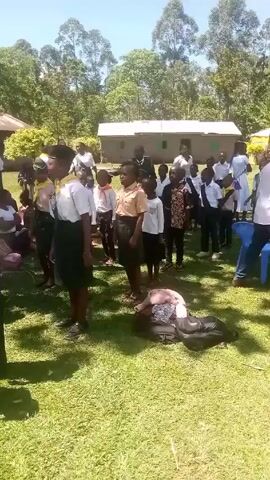 Here is a concise description of the video:

The video depicts a group of children, primarily dressed in school uniforms of varying styles, standing on a grassy lawn outdoors. Some of the children are wearing scout-like uniforms with scarves. There are also a few adults present among them. One person, possibly a child, is lying on the ground with their head resting on a backpack. In the background, there is a building and trees. The overall setting appears to be a school or community gathering. The lighting indicates it's daytime.