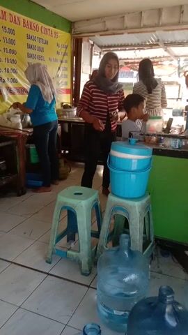 A woman wearing a headscarf and a striped shirt is sitting on a stool in front of a dispenser. A child is standing next to her, reaching towards the dispenser. There are several large water jugs in the foreground and background. A sign with text in an Asian language is visible on the left. The scene appears to be an outdoor food stall or shop.