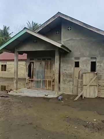 The video shows a cloudy, overcast sky above a village scene. The foreground is dominated by muddy, uneven terrain with scattered debris, including plastic bottles and what appears to be a broken pipe. Several unfinished or dilapidated buildings are visible. On the left, a concrete house with a gray stucco finish and a sloping roof is partially obscured by a smaller structure with a corrugated metal roof. The main house has a wooden door and several windows, some boarded up. Adjacent to this house, a taller building with a tin roof and wooden stilts stands. Further to the right, another concrete building with large, open window frames and a rusted metal roof is prominent. The overall impression is one of disrepair and abandonment, possibly due to natural disaster or ongoing construction. No people are clearly visible in the footage.