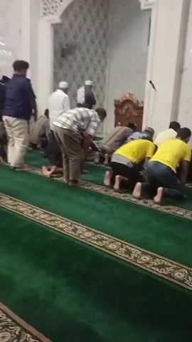 A group of men are standing and kneeling on a green carpet in what appears to be a mosque. They are dressed in a variety of clothing, including shirts, pants, and traditional Islamic attire. Some of the men are wearing skullcaps. The men are facing the same direction, and some are bowing their heads. The walls of the room are white and decorated with geometric patterns. There is a large ornate chandelier hanging from the ceiling. The lighting is bright and even.