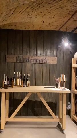 A wide shot captures the corner of a dimly lit wine cellar, revealing two walls with a brown, grooved wooden texture. On the left, a long, light wooden table stands with a display of bottled products, presumably wine, on top. A wooden sign above the table reads "Valmiermuiža". Adjacent to the table, a shelf unit holds several wooden wine barrels, some of which bear a star logo. The cellar's ceiling is made of rough, light brown material, and a track light with two spotlights illuminates the space.