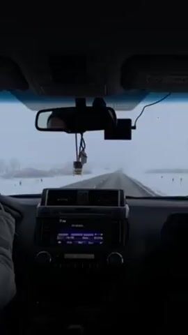The video shows the dashboard view from inside a car driving on a long, straight road covered with snow. The weather is foggy or snowy, obscuring the view ahead. The car's radio is on and tuned to 101.80 MHz.  A rear-view mirror hangs from the top of the windshield, and there is a dash cam attached as well.