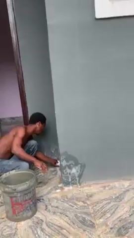 A shirtless man is cleaning the corner of an exterior wall. He is crouching next to a bucket and using a sponge to wash a dark-grey painted wall that has visible white stains. The wall is adjacent to a doorway with a purple wall inside. The floor is covered in patterned grey and brown tiles. A bottle of soda is visible on the floor in the background.
