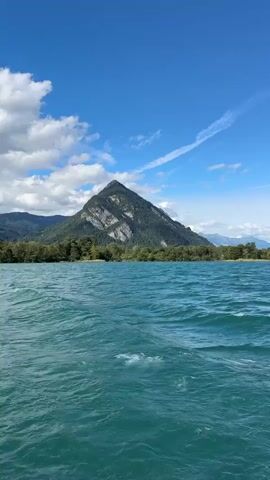 The video is a low-angle shot from a moving boat on a bright, sunny day. The water is a vibrant turquoise and is choppy with small waves. In the distance, a tree-lined shore separates the water from a majestic, conical mountain with grey rock faces and green slopes. The sky is a brilliant blue with scattered white clouds. The camera pans slowly from left to right, revealing more of the expansive mountain range in the background. There are no people or man-made objects visible in the frame.