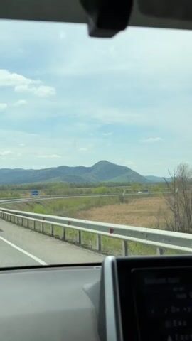 The video shows a view from the driver's seat of a car traveling on a highway. A large truck passes on the left. In the distance, a green mountain dominates the landscape under a sky with sparse clouds. The dashboard of the car is visible, featuring a digital display. Music is playing in the car.
