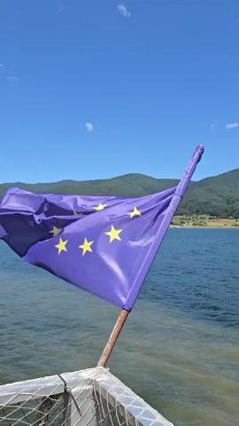A European Union flag flies on a boat, with a large body of water and green hills in the background. The sky is clear and blue. The camera pans across the water.