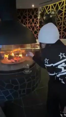 A woman in a white beanie and a black sweater with white stars is roasting a marshmallow over a fire in an enclosed fireplace. The fireplace has a black metal hood and a base covered in black and grey tiles. A glass teacup is sitting on the fireplace base. The background features decorative wooden panels with a geometric pattern. The lighting is warm and inviting.