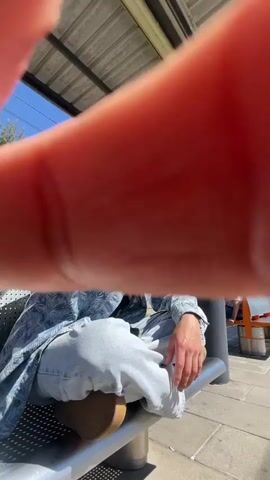A young man with black hair and sunglasses sits cross-legged on a metal bench at a train station. He wears a patterned blue shirt over a black t-shirt and light gray jeans with brown shoes. The camera starts close to his outstretched hand, then pulls back to show him gazing towards the sunlight with his hands clasped. In the background, another person sits on an orange bench. The station has a metal roof supported by a black column, and the sky is bright blue with trees visible in the distance.