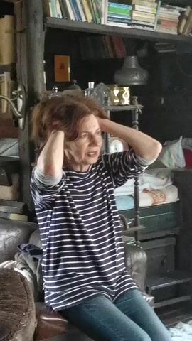 A woman with brown hair in a striped long-sleeved shirt and blue jeans sits in a room and holds her hands to her head. She expresses pain. Another woman with brown hair wearing a black short-sleeved shirt walks past. The camera pans to a window with a view of a lake and cliffs.
