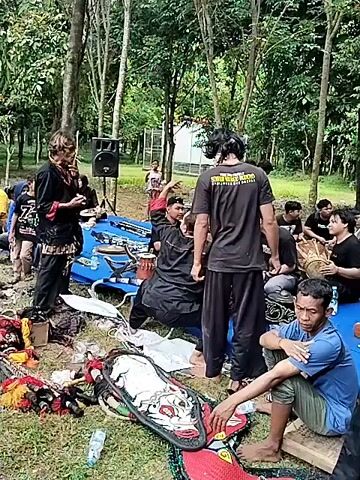 The scene is a wooded area with a group of people gathered, likely preparing for a performance. The environment is a grassy clearing with tall, thin trees surrounding it.

Several individuals are seated on a blue tarp, some drumming on traditional instruments. Others are standing or moving around. One person is dressed in black, and may be preparing to perform some ritual. The ground is littered with various props and equipment, including masks and decorative items. A speaker stands near the edge of the tarp, suggesting music or amplified sound will be part of the event.

The atmosphere appears to be one of anticipation and preparation. The people are interacting, possibly coordinating before a performance.