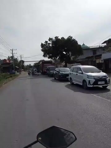 The video shows a street scene from the perspective of someone riding a motorcycle. The street is paved and appears to be a two-lane road. There are several vehicles present, including cars, trucks, and motorcycles. On the left side of the road, there are some buildings and utility poles. On the right side, there are more buildings and trees. The sky is overcast. The video captures a moderate amount of traffic, with vehicles moving in both directions. One motorcycle in the foreground has its side mirror visible. A tricycle is seen carrying wooden planks. A black SUV is also visible on the right side of the road. The scene suggests a typical day in a town or city with a busy road.