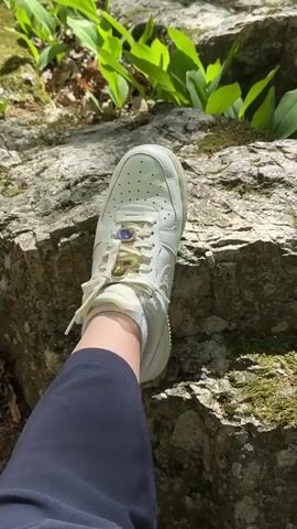 A close-up shot shows a person's leg wearing a white sneaker with decorative jewelry on the laces resting on a moss-covered rock. The person is wearing dark blue pants. Green plants are visible in the background. The scene appears to be outdoors in a natural setting.