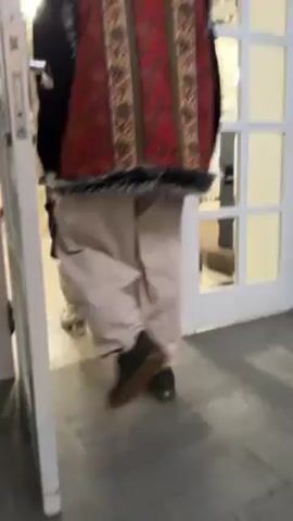 A person wearing a red patterned vest, light-colored baggy pants, and dark shoes walks through a doorway. The doorway leads into a room decorated with Christmas lights and greenery. A white door is partially open on the left of the frame. The floor visible in the foreground is dark gray.