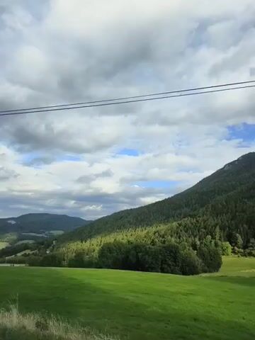 The video captures a scenic landscape from a moving vehicle. The viewpoint is from the passenger side, as evidenced by the partially visible car door in the lower left corner. The scene is dominated by rolling green hills and mountains covered in lush green trees. A wide, open grassy field is in the foreground, with a paved road visible at the very bottom. In the distance, a valley stretches out with more hills and what appears to be a road winding through it. The sky is a mixture of blue with dramatic, puffy white clouds. Two thin, dark power lines run horizontally across the top portion of the frame, suggesting the presence of infrastructure in the rural setting. The lighting indicates it is daytime, likely late afternoon or early evening, with some areas in shadow and others illuminated by sunlight. There are no people visible in the video. The overall impression is one of natural beauty and tranquility in a mountainous region.