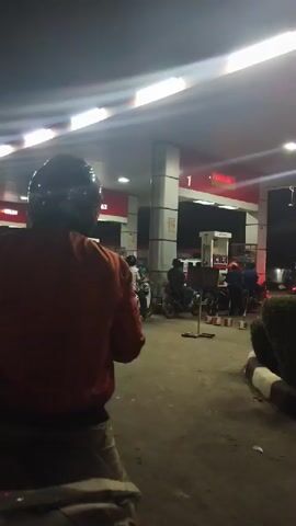 The video shows a perspective from a person riding a motorcycle at a gas station at night. The person in the foreground is wearing an orange jacket and a helmet, with their legs visible. Other motorcycles and people are present at the gas station. The gas station has illuminated signage and white pillars. The ground is paved. The scene appears to be a typical nighttime scene at a gas station with customers.