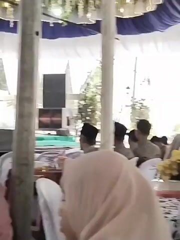The video shows an outdoor event under a tent. There are people seated at tables, and some are standing. A woman wearing a light pink hijab is in the foreground, partially obscuring the view. Another woman in a blue hijab is seated to her left. In the background, several people are standing, some wearing hats. Large speaker cabinets are visible behind the standing individuals. The tent is decorated with white and blue fabric. The overall impression is of a gathering, possibly a wedding or a celebration.