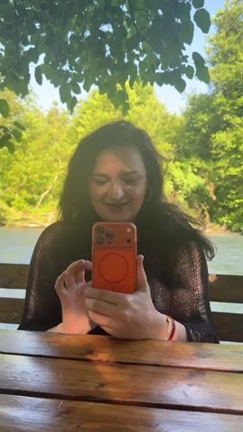 A medium shot video shows a young woman sitting at a wooden table outdoors. She has long dark curly hair and is wearing a black sheer top. The woman is holding an orange smartphone in her hands and looking at the screen. Behind her, a river is visible, and the background is filled with lush green trees and foliage. The lighting suggests it is daytime, with sunlight filtering through the leaves overhead, casting dappled shadows on the scene. The woman smiles and waves at the camera at one point.
