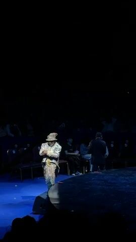 A person in a patterned jacket and hat stands on a blue stage with their back to the camera, facing another person holding a green balloon. The second person pops the balloon, creating a cloud of smoke around their head. An audience is seated in the dark background.