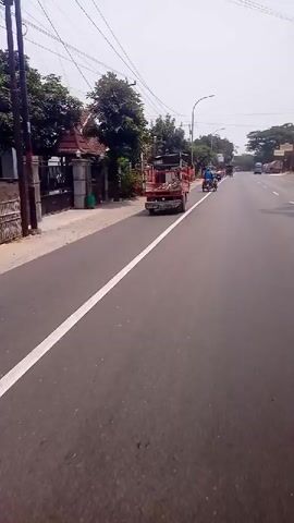 The video shows a view from the perspective of someone riding a motorcycle on a paved road. The rider is traveling in the right lane, with a white line marking the edge of the lane. To the left of the road, there is a sidewalk and a residential area with houses, fences, and trees. A red truck with an open cargo bed is in front of the motorcycle, also traveling in the right lane. Further down the road, there are other vehicles, including motorcycles and trucks, and trees lining the sides of the road. The sky is overcast.