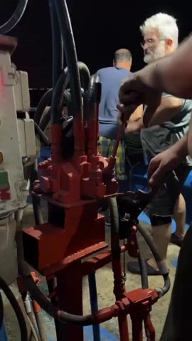 A medium shot captures a group of men operating a piece of heavy machinery on what appears to be a boat at night. In the foreground, a red hydraulic valve assembly with multiple hoses connected is prominently displayed. A man with gray hair and a beard, wearing glasses and a yellow shirt, is in the process of operating a lever on the valve assembly. His hands are visible, and he is looking towards the machinery. To his left, another man in a blue shirt and checkered shorts is also facing the machinery, his back partially to the camera. A third person, whose hands are visible on a different lever to the right, is partially obscured. In the background, a control panel with several illuminated red lights and switches is visible, mounted on a blue structure. The overall environment is dimly lit, suggesting it is nighttime, with the lighting focused on the machinery and the individuals working with it.