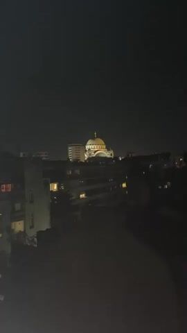 A nighttime cityscape is captured in a video, focusing on a large, illuminated building with a golden dome topped by a cross. The building appears to be a church or cathedral. Surrounding it are lower-rise apartment buildings, some with windows glowing with interior light. The sky is dark and overcast, and the overall impression is one of a peaceful, urban night.