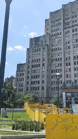 The video shows a tall, gray building with many windows on a sunny day. The building appears to be several stories high and has a boxy, tiered design. There are some palm trees and other greenery in front of the building. There's a yellow barrier in the foreground with some graffiti on it. A street light is also visible. The camera tilts upwards to show the full height of the building. The sky is blue with some white clouds.