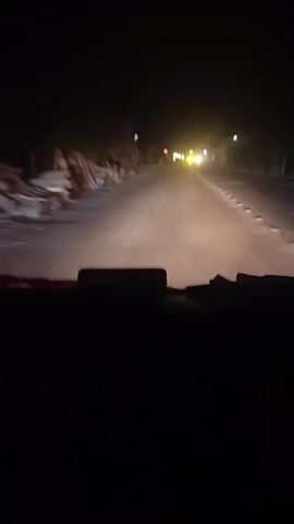 The camera is positioned inside a vehicle driving down a dark, paved road at night. The headlights illuminate the road ahead, revealing a worn surface with white road markings. Buildings and vegetation line the sides of the road, but details are obscured by the darkness. Distant lights from other vehicles and street lamps are visible further down the road. The dashboard is visible at the bottom of the frame, appearing dark and undefined.