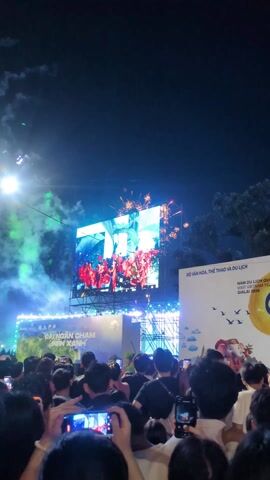 The video shows a nighttime scene of a large crowd gathered to watch fireworks. The fireworks are being launched behind a large screen displaying a performance. A large sign on the right side of the screen reads “Bộ Văn Hóa, Thể Thao và Du Lịch” and “Năm Du Lịch Quốc Gia Việt Nam YÊN BÁI GIA LAI 2026”. Many members of the crowd are holding up their phones to record the event. There are spotlights and stage lights illuminating the area. The atmosphere appears celebratory.