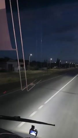 This video captures a nighttime drive on a highway near Kokpekti, Kazakhstan. The perspective is from inside a vehicle, likely a bus or truck, looking forward through the windshield. The headlights of the vehicle illuminate the asphalt road, which has a prominent white line dividing the lanes.

The scene is set at dusk or early night, with a dark, cloudy sky overhead. Streetlights line the side of the road, casting pools of light and creating a sense of an approaching settlement or town. In the distance, other vehicles can be seen with their headlights on, traveling in the same direction. The surrounding landscape appears rural, with low-lying buildings and vegetation visible on the left side of the road.

A dashcam is mounted on the dashboard in the foreground, displaying a small screen. Some thin, pink-colored wires or cables are also visible hanging from the upper left of the frame, possibly part of the vehicle's interior or equipment. The overall mood is one of travel and transition, with the ongoing drive through the dim light suggesting a journey. There is no discernible text in the video.