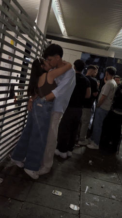 Couple kisses in alleyway while group gathers nearby