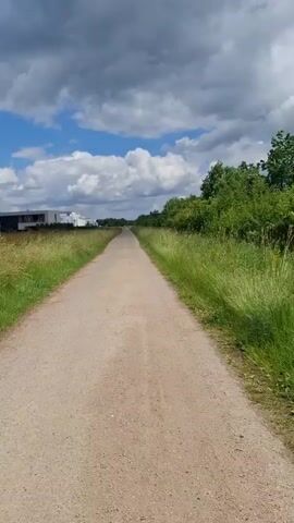 A dirt path winds through a grassy field under a partly cloudy sky. On the left, a modern building with a dark roof is visible in the distance. On the right, lush green trees line the path. The camera pans slowly, showing the changing scenery as the viewer moves along the path.