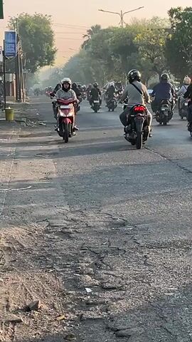 A group of people on motorcycles are riding down a street with a lot of potholes. The sun is low in the sky, casting a warm glow. There are trees and buildings on either side of the street. One person in the foreground is wearing a gray hoodie and a white helmet. Another person behind them is wearing a pink hijab. The road is made of asphalt and is in poor condition.