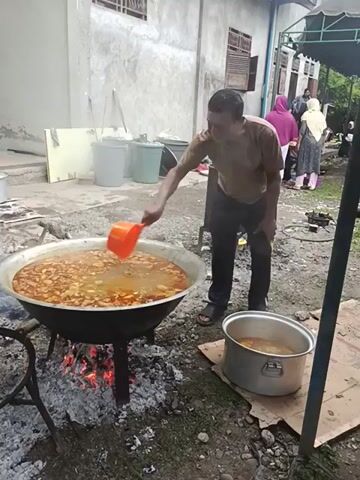 A man is pouring liquid from a metal pot into a very large pot of stew being cooked over an open fire outdoors. The man is wearing a tan shirt, dark jeans, and black sandals. He is standing in a yard that is partially paved with gravel and partially covered with grass. There are several people under a white tent nearby. A second metal pot sits next to the large cooking pot. Three light-colored buckets are lined up near a white building with closed windows. The man is using a large orange ladle to scoop the liquid from the metal pot. The large pot is supported by a metal tripod over the embers of the fire.